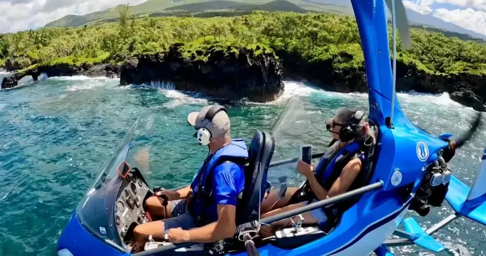 Pilot flying autogyro over Maui coastline with clear blue skies